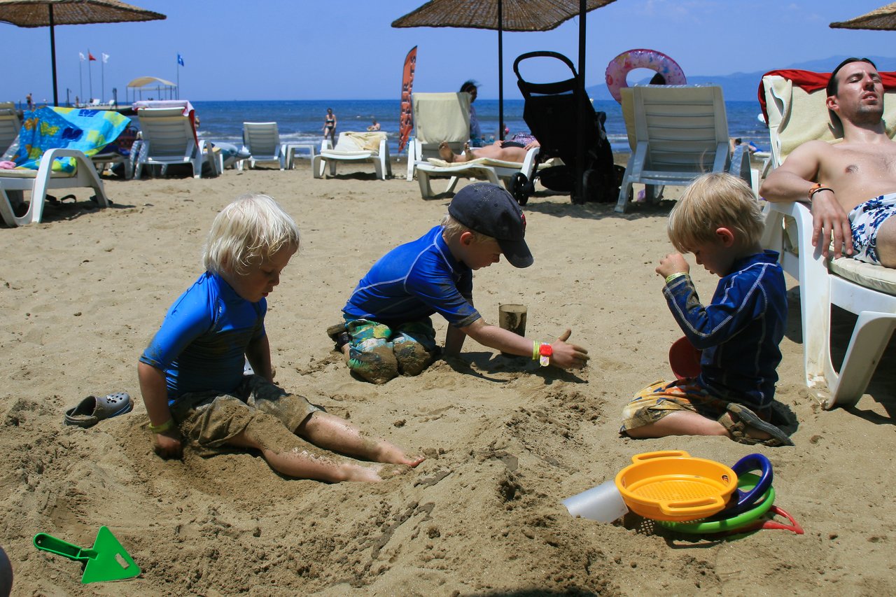 Three children in swimwear play in the sand on a beach, using toys to dig and build.