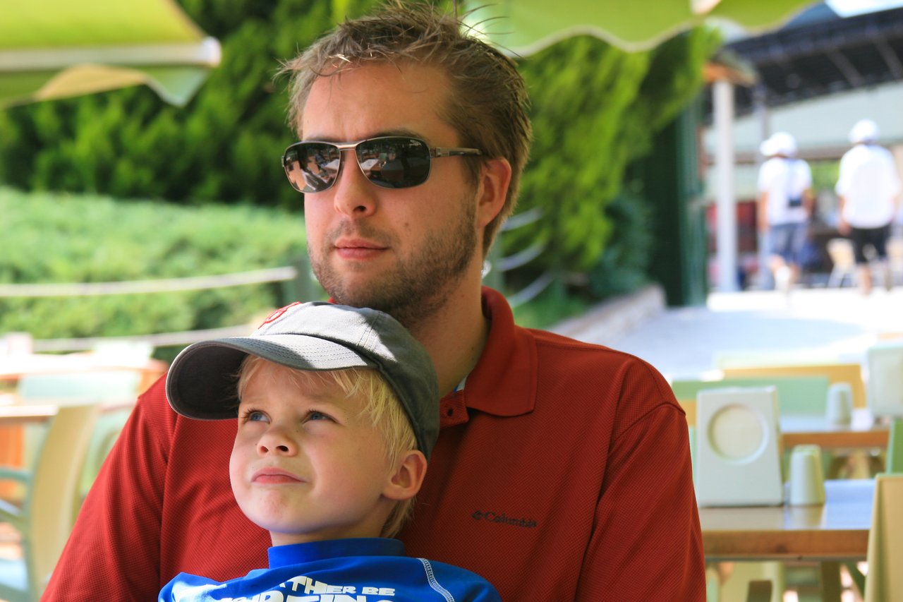 A man wearing sunglasses and a red shirt sits with a young boy in a cap on his lap.