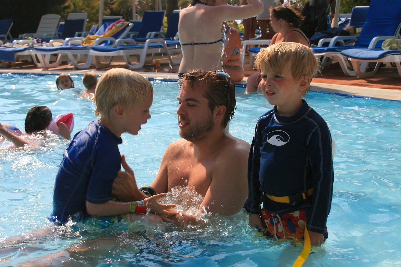 A man in a pool holds a young child while another child stands nearby, both wearing swim shirts.