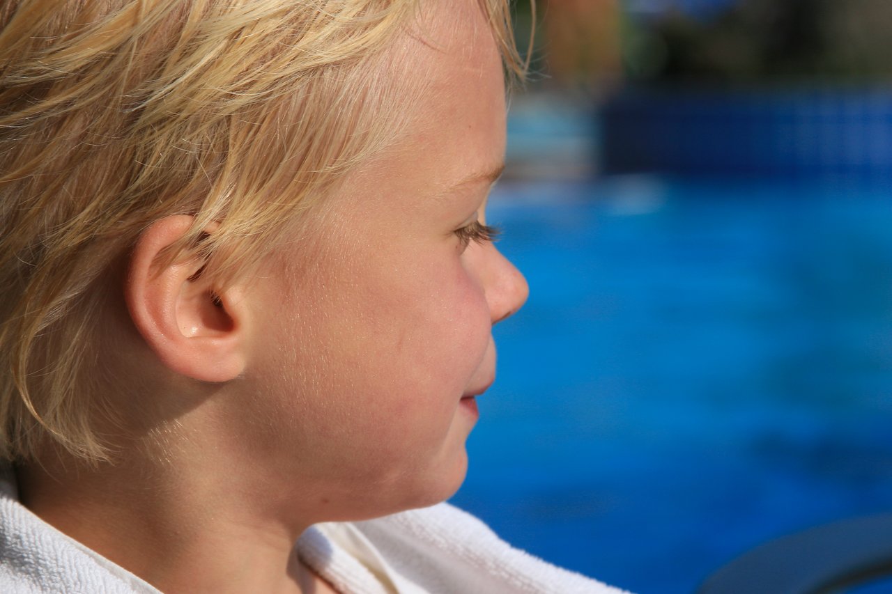 A child with wet blonde hair sits near a swimming pool, wrapped in a white towel.
