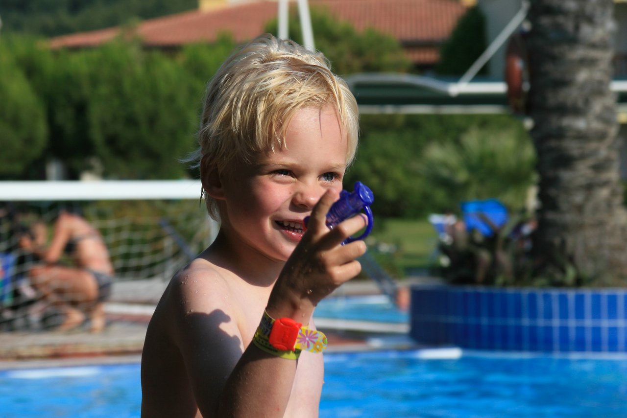 A smiling child by the pool holds a small purple water toy, appearing to aim it playfully.