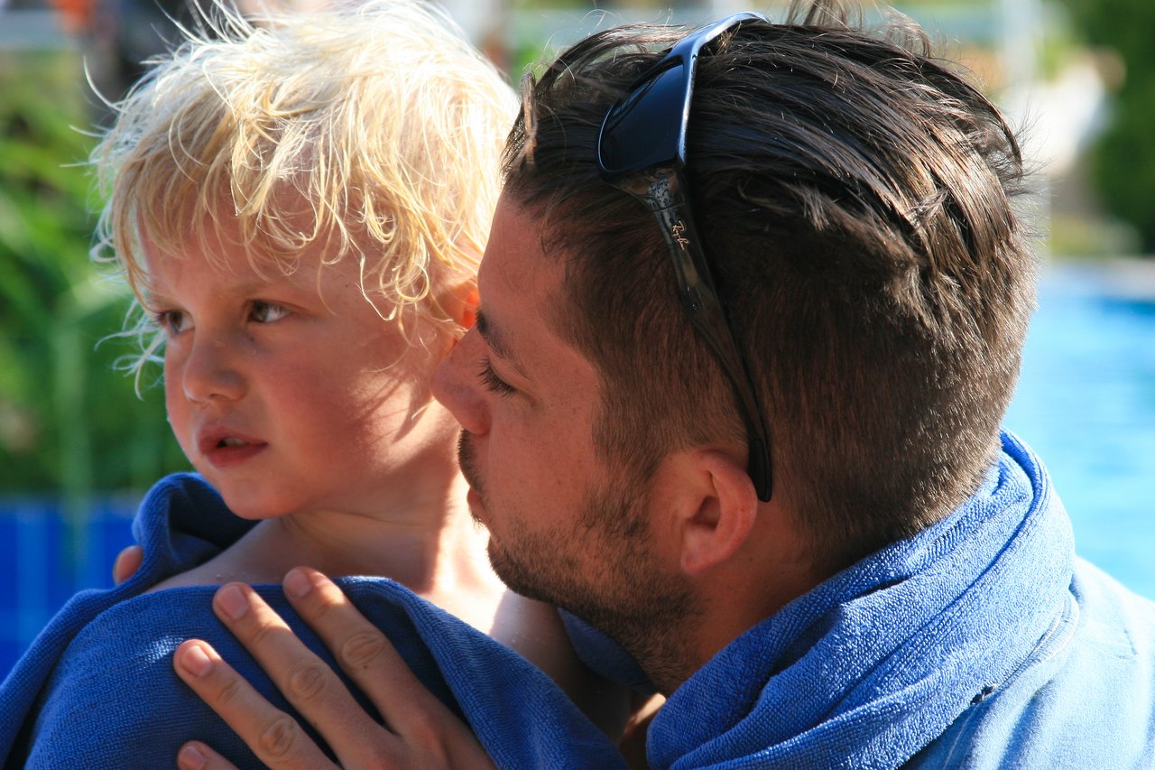A man wrapped in a blue towel holds a young child close, speaking softly near a swimming pool.