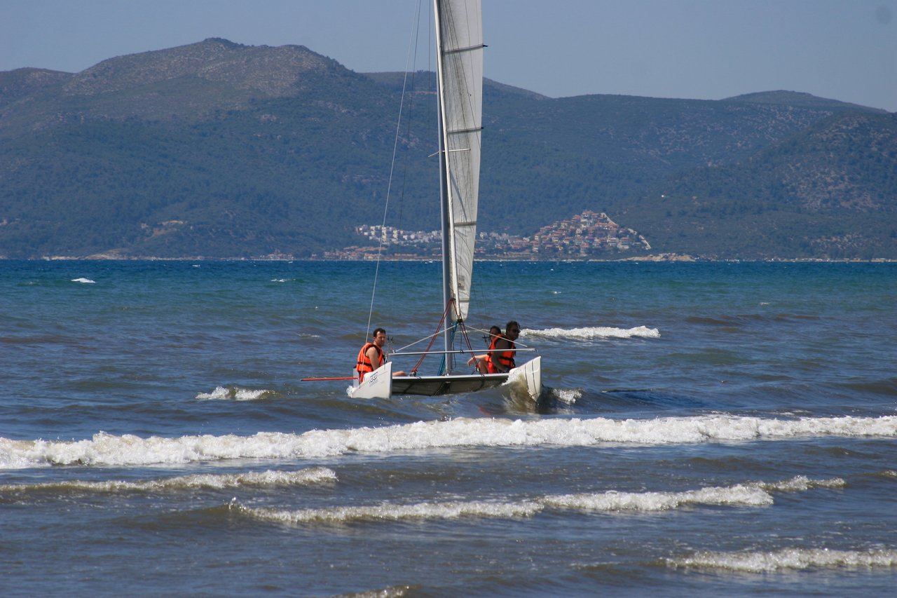 Two people wearing life jackets sail a small boat through shallow waves near the shore.