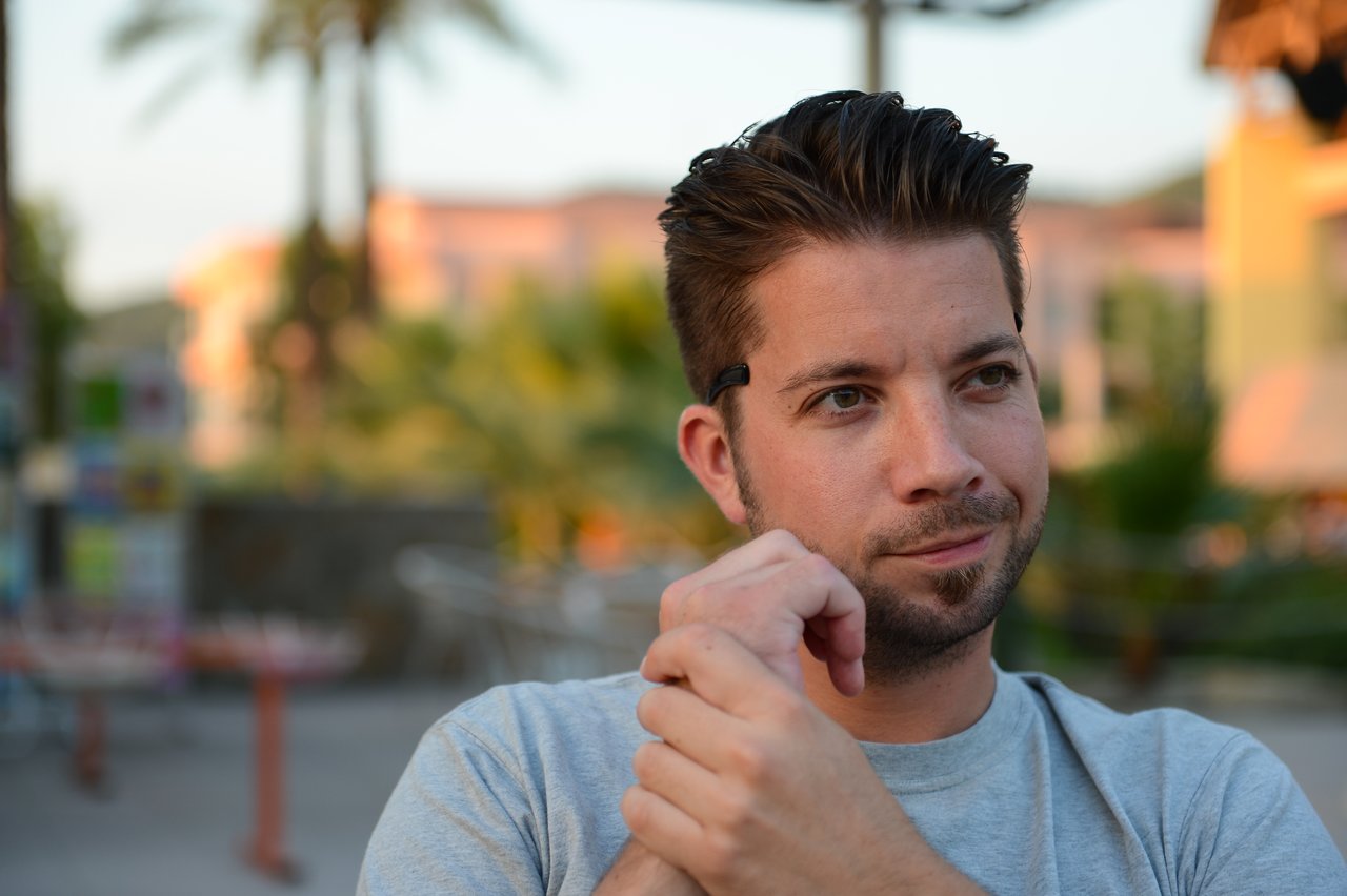 A man with short dark hair and a beard sits outside, wearing a gray shirt and a small earpiece.