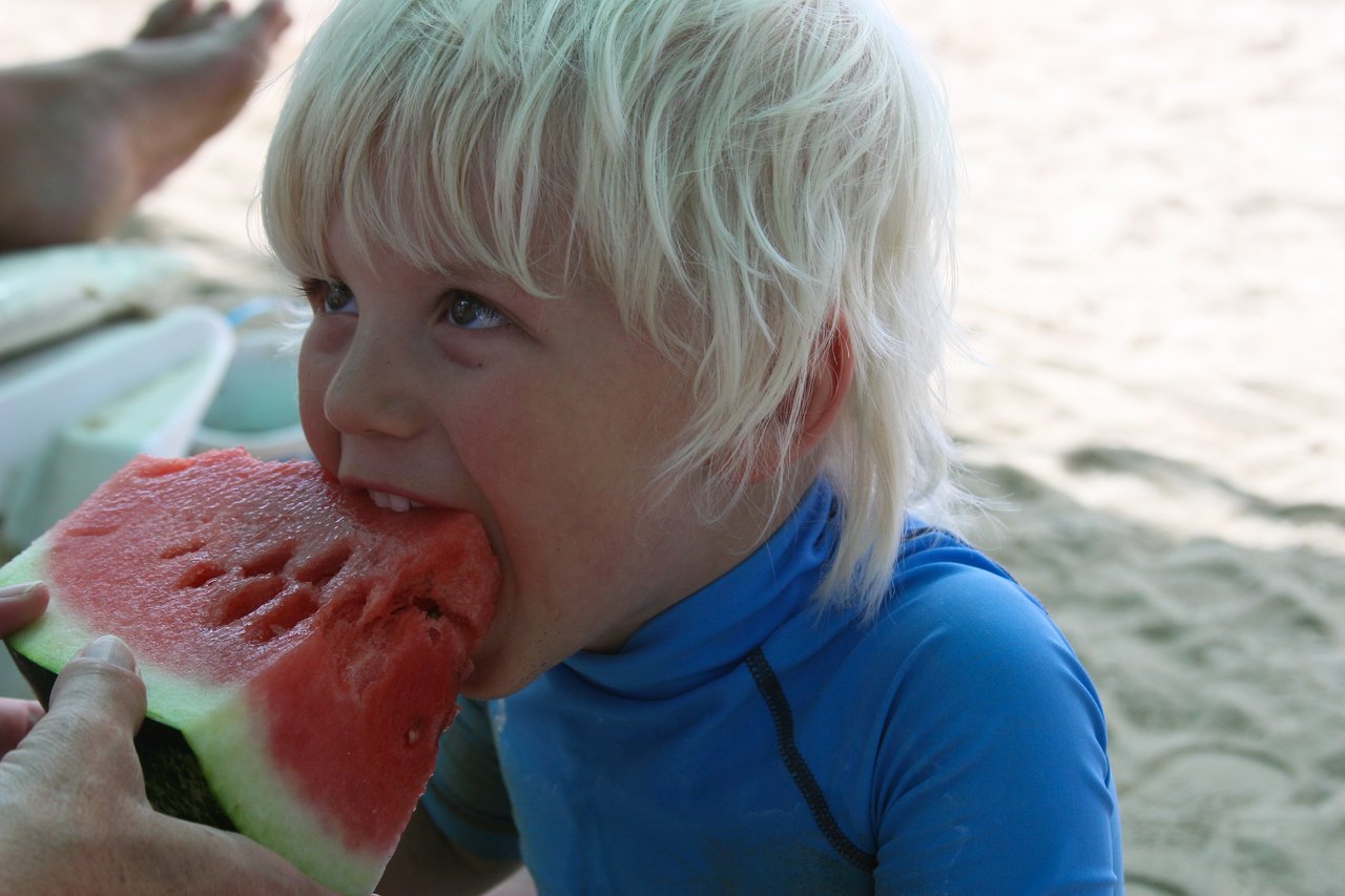 A young child in a blue shirt takes a big bite of a watermelon slice on a sandy beach.