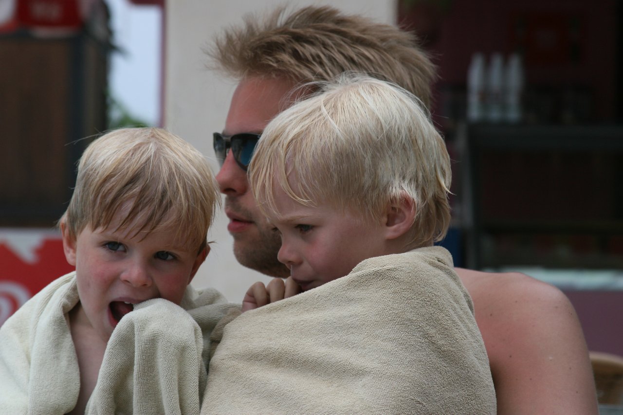 A man wearing sunglasses sits with two young children wrapped in towels, possibly after swimming.