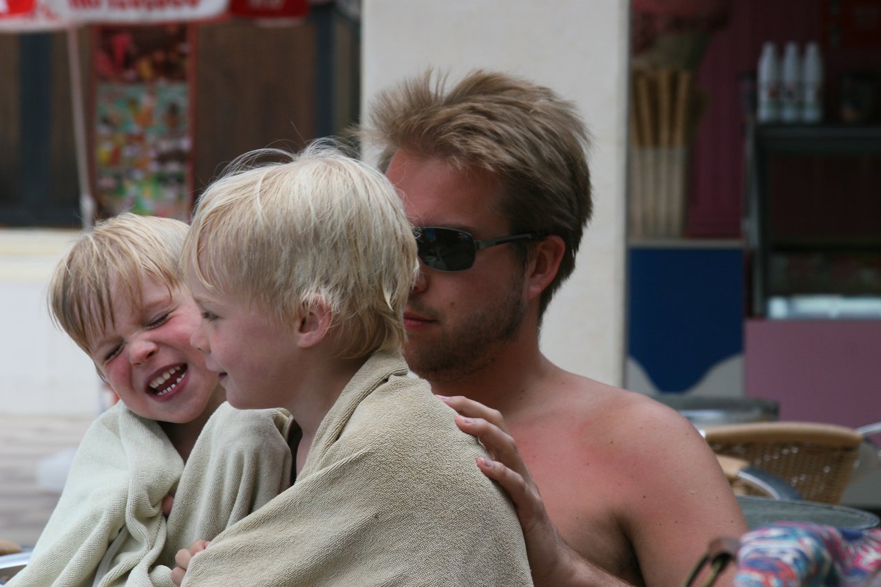 A man wearing sunglasses sits with two children wrapped in towels, one smiling and the other laughing.