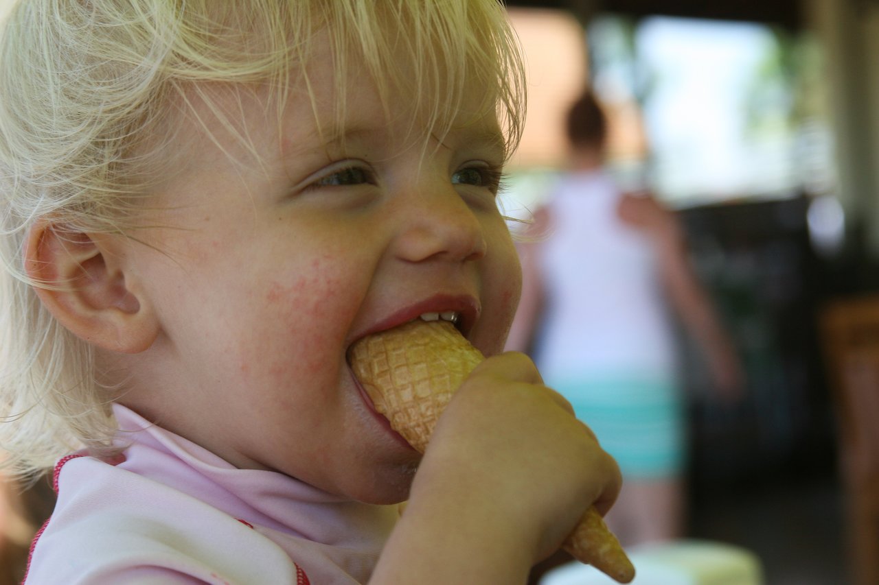 A young child with blonde hair bites into an ice cream cone, smiling.