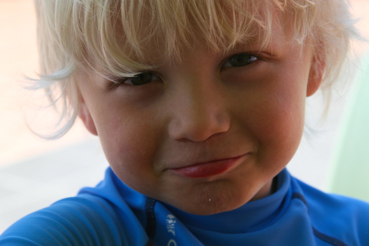 A young child with blonde hair and a blue shirt looks directly at the camera with a slight smile.