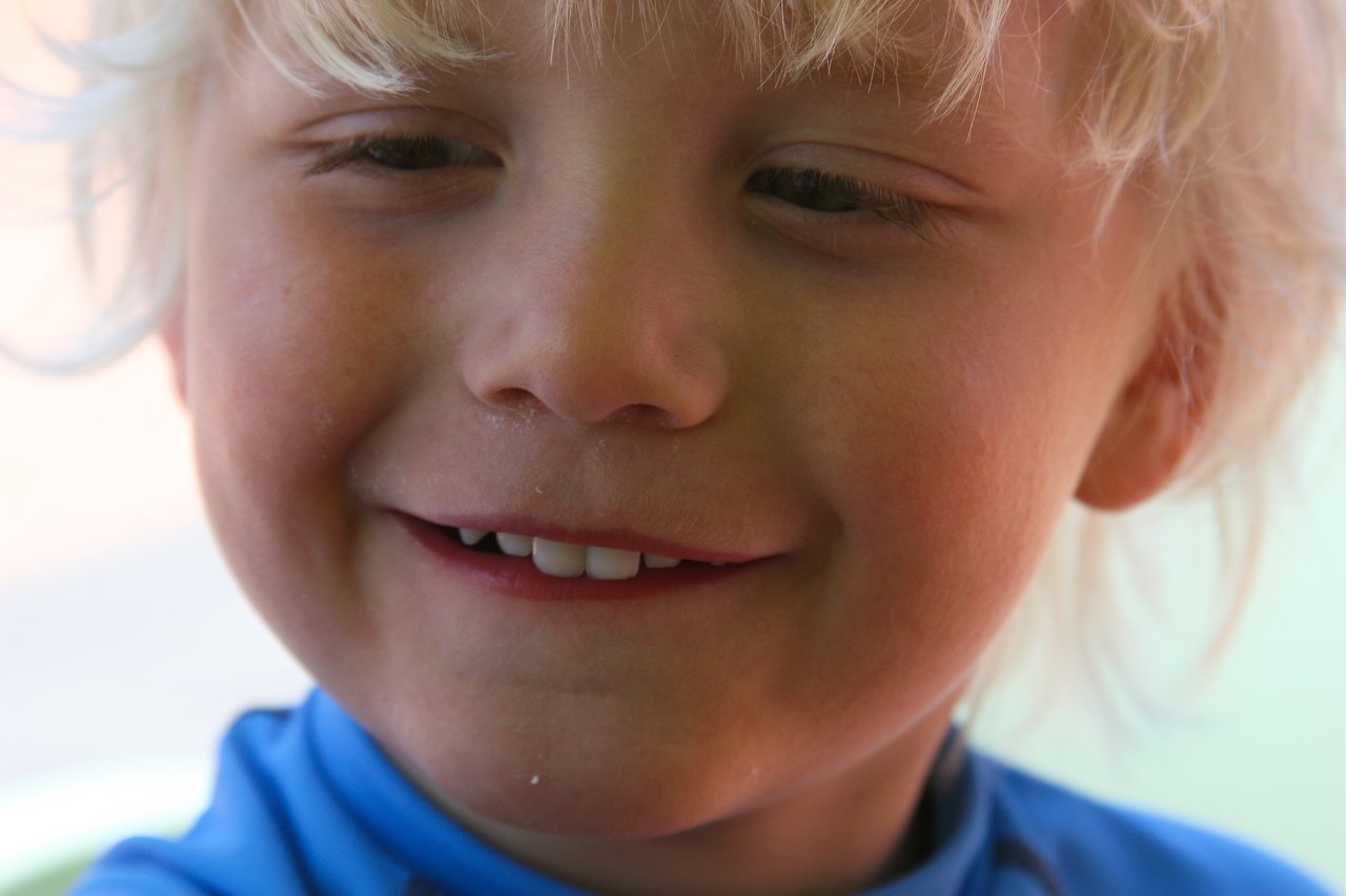 Close-up of a smiling child with blonde hair, wearing a blue shirt.