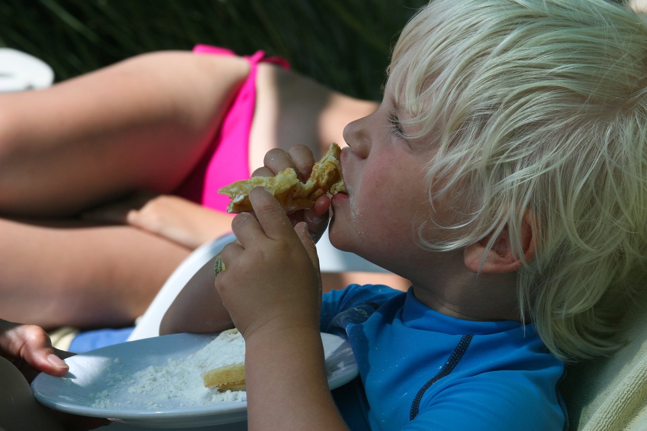 A young child with blonde hair eats a piece of food while holding a plate with more food.