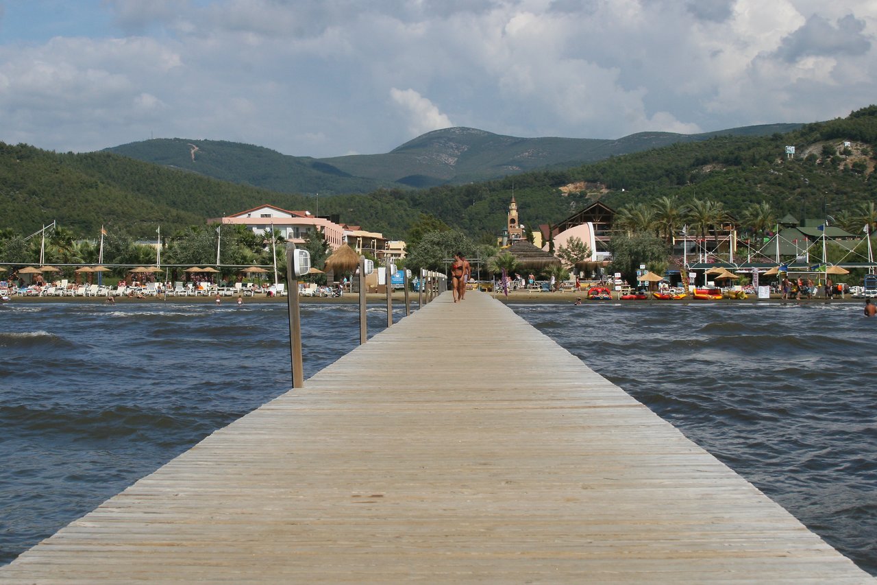 A person walks on a wooden pier extending over the water toward a busy beach with buildings and mountains.