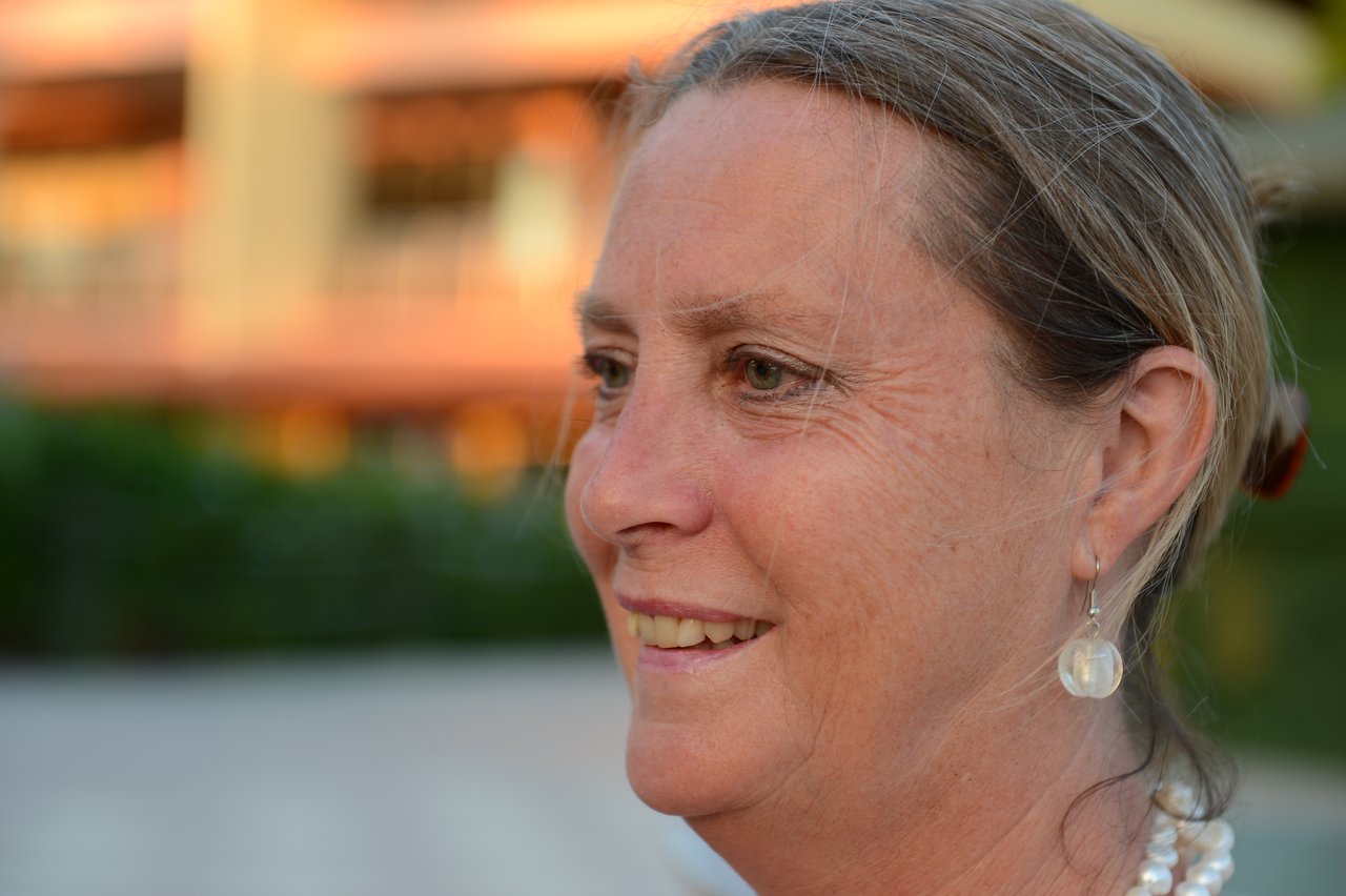 A woman with light skin and pearl earrings smiles while looking to the side, with a blurred background.