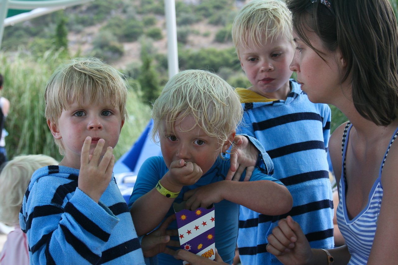 Three young children in blue striped towels eat snacks while a woman in a striped swimsuit interacts with them.