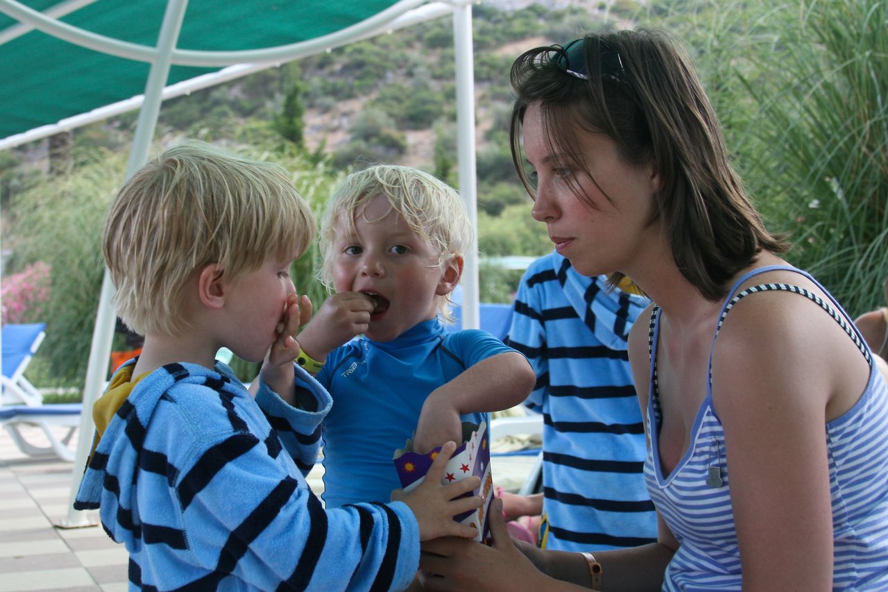 A woman and two children in striped clothing share a snack outdoors under a shaded area.