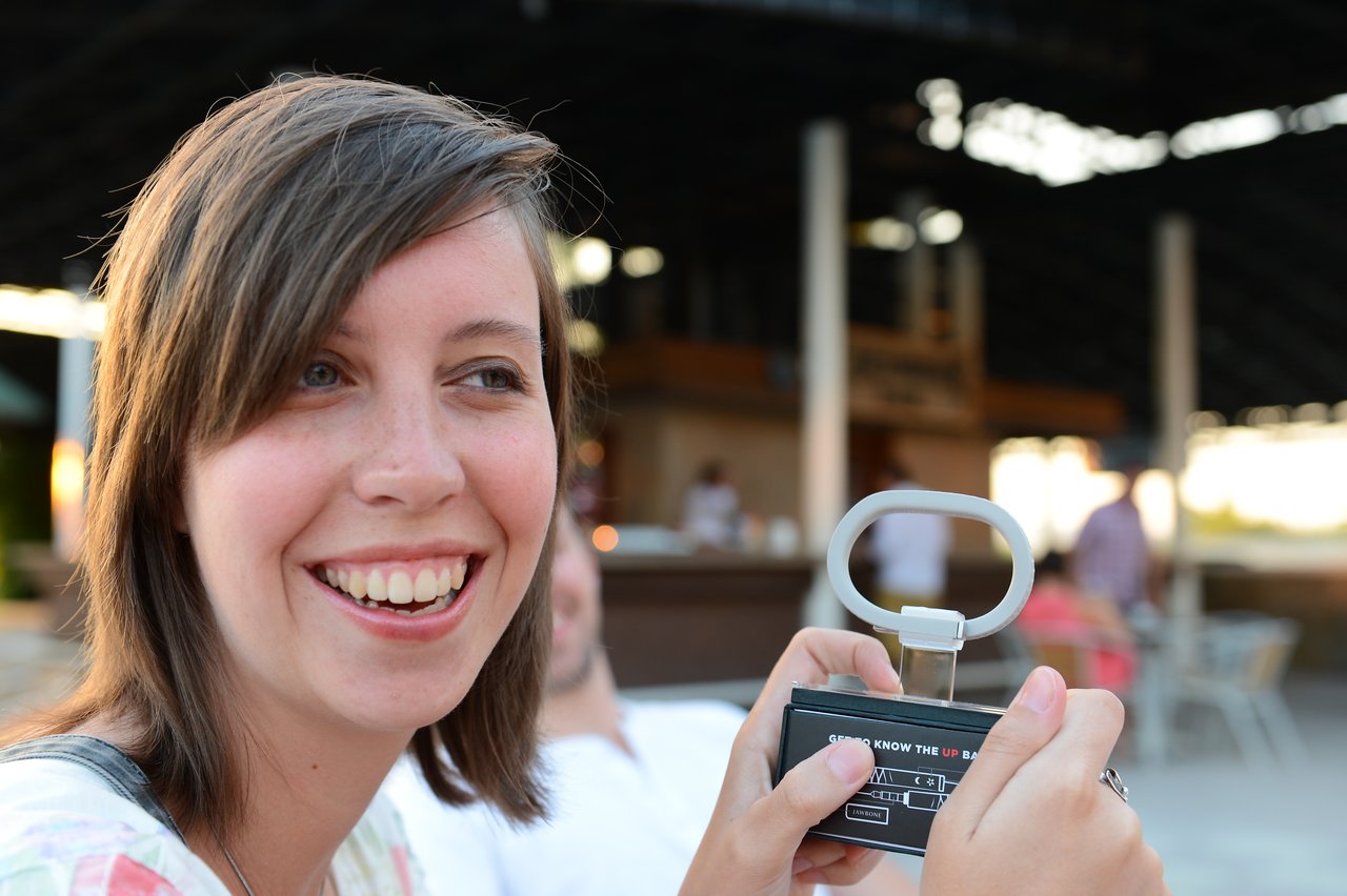 A smiling woman holds a small handheld device with a loop, possibly a game or gadget, in an outdoor setting.