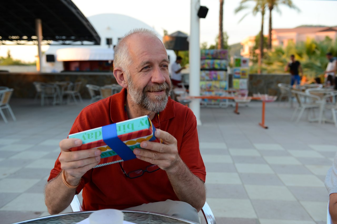 A man in a red shirt smiles while holding a wrapped birthday gift with a blue ribbon.