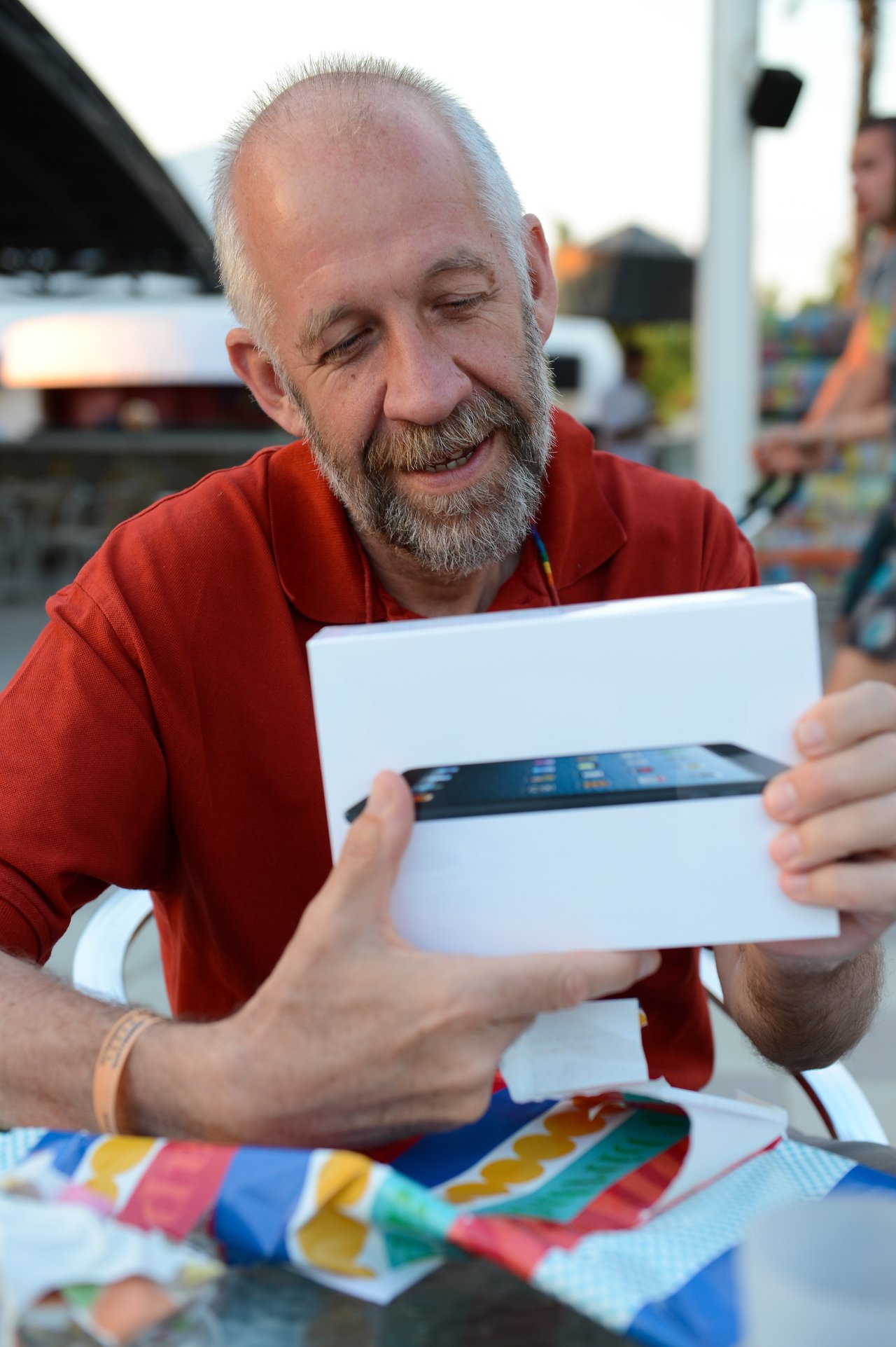 A man in a red shirt smiles while holding a new tablet box at a table with gift wrapping.