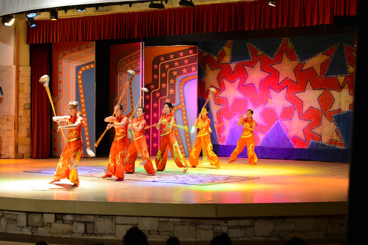 A group of performers in orange outfits spin batons on a brightly lit stage with a colorful backdrop.