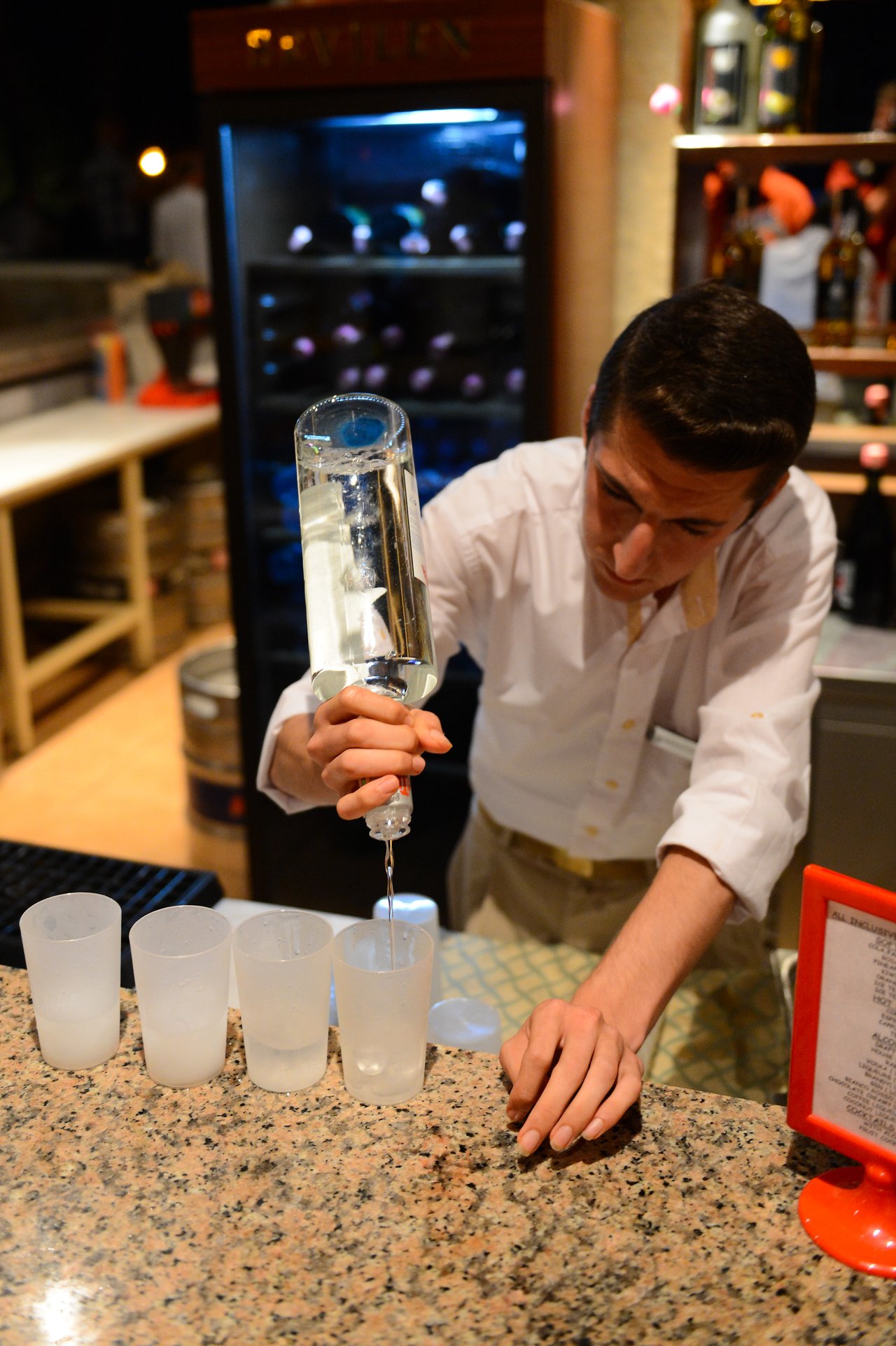 A bartender pours a clear liquid into plastic cups at a bar counter.