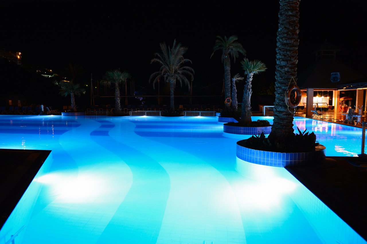 A large swimming pool glows with blue lights at night, surrounded by palm trees and a nearby seating area.