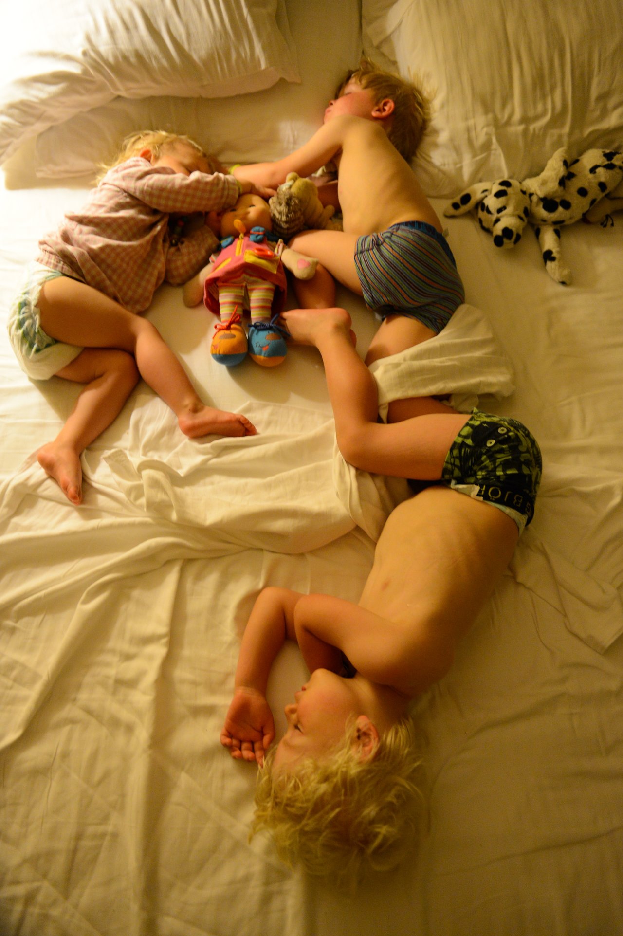 Three young children sleep on a bed, holding stuffed toys and dolls, with blankets and pillows around them.