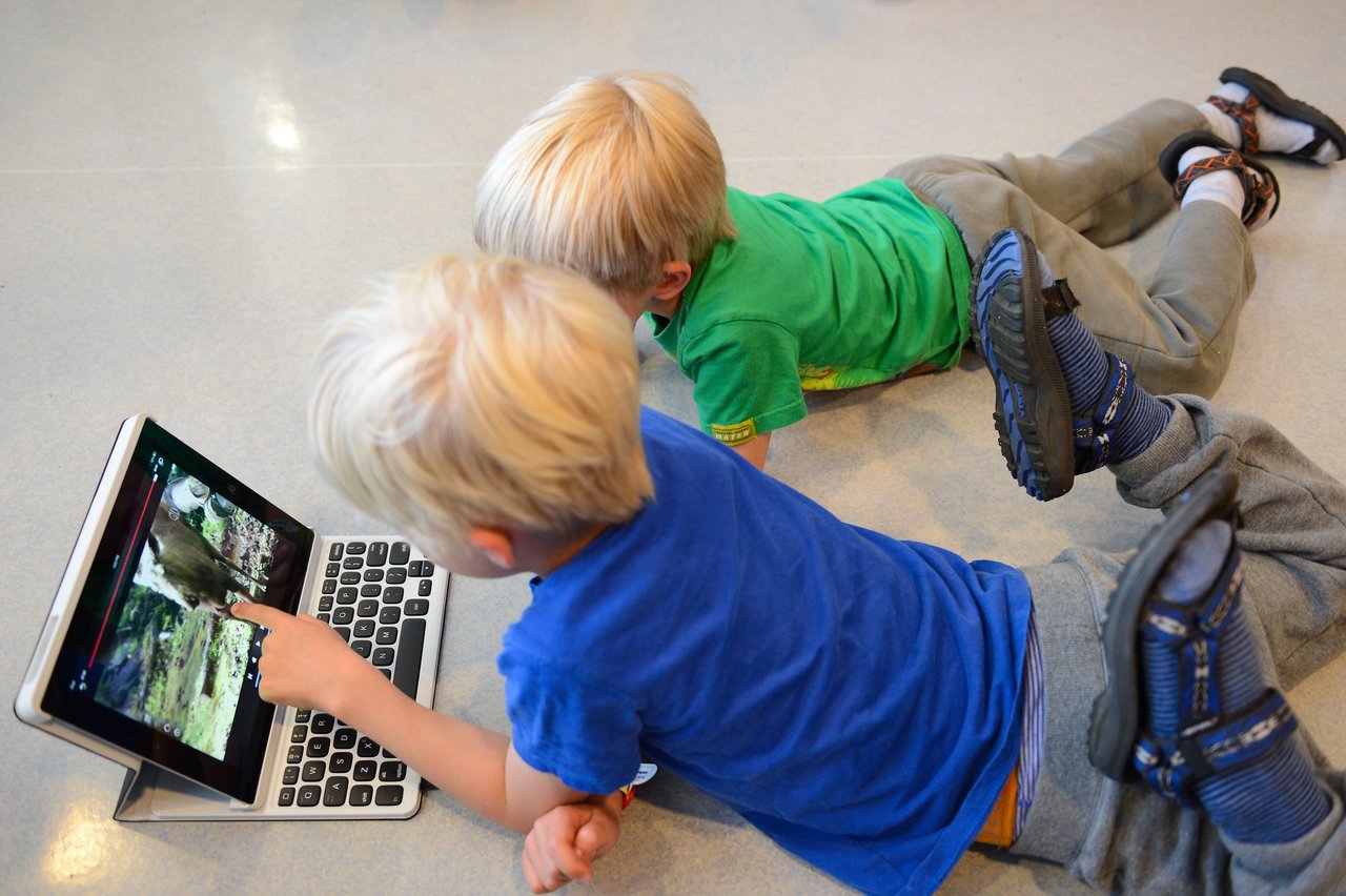 Two children lie on the floor watching a video on a tablet, with one pointing at the screen.