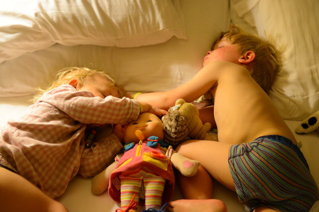 Two young children are sleeping on a bed, holding stuffed toys and a doll.