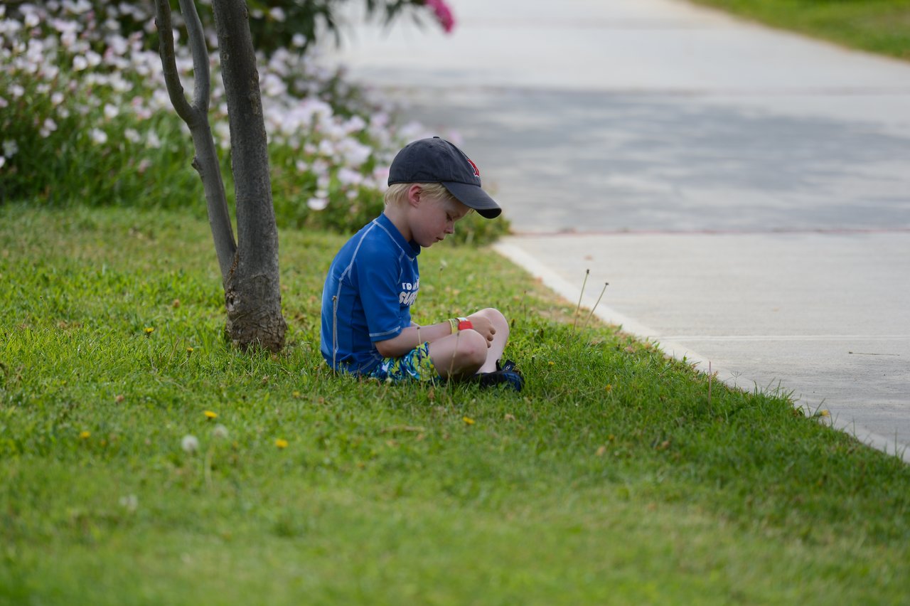 A young boy in a blue shirt and cap sits on the grass, focused on an object in his hands.