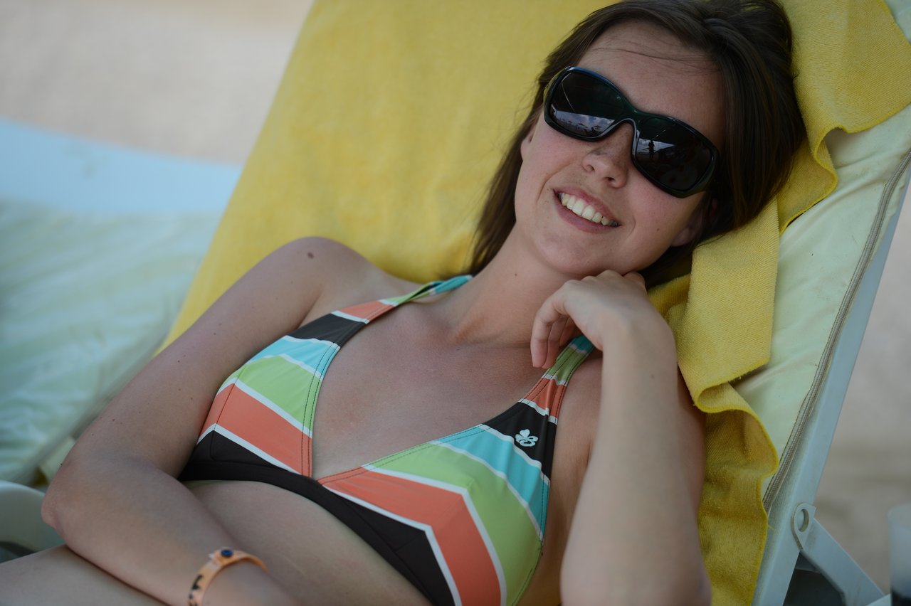 A woman wearing sunglasses and a striped bikini smiles while reclining on a lounge chair with a yellow towel.