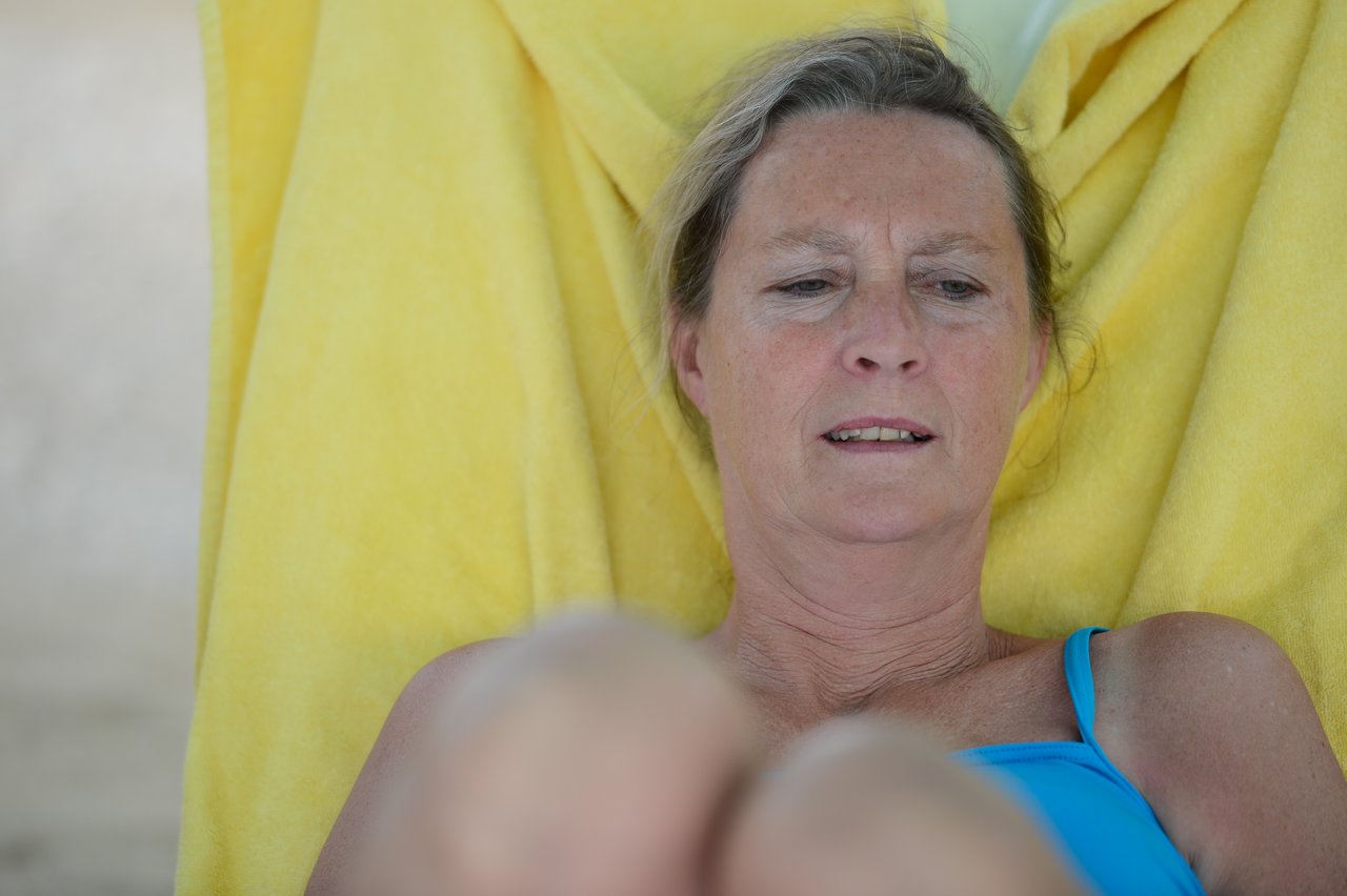 A woman in a blue swimsuit reclines on a chair with a yellow towel, looking down with a neutral expression.