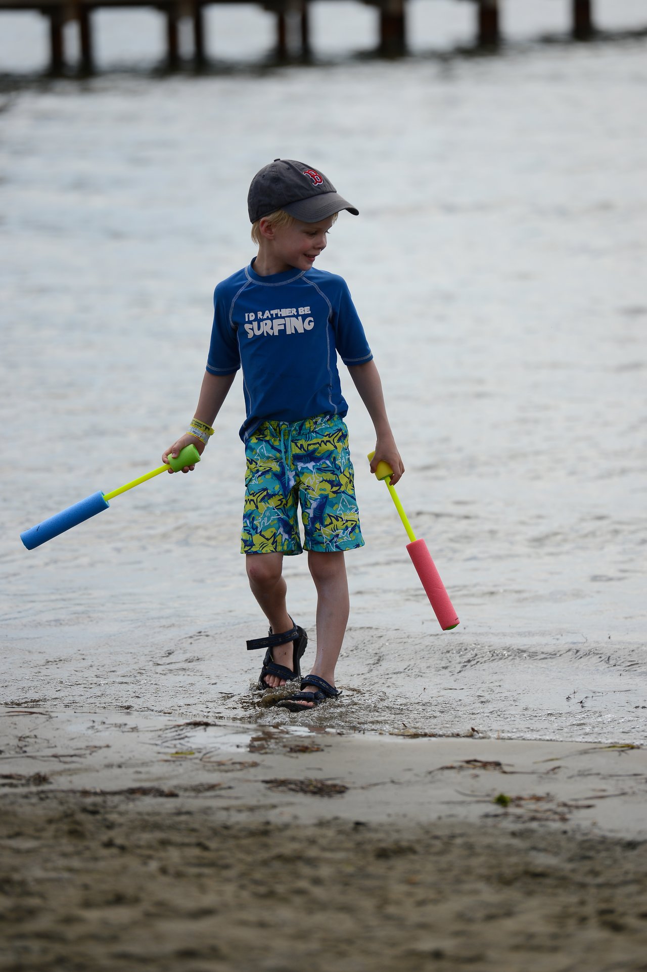 A young boy in a blue shirt and cap walks on the beach holding two foam water toys.