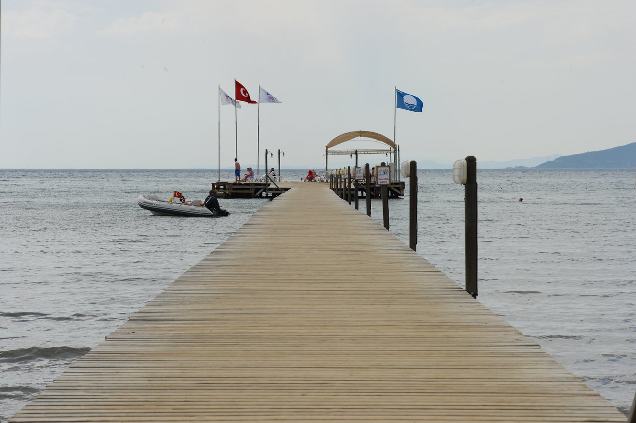 A wooden pier extends into the sea, leading to a dock with flags and a small boat nearby.