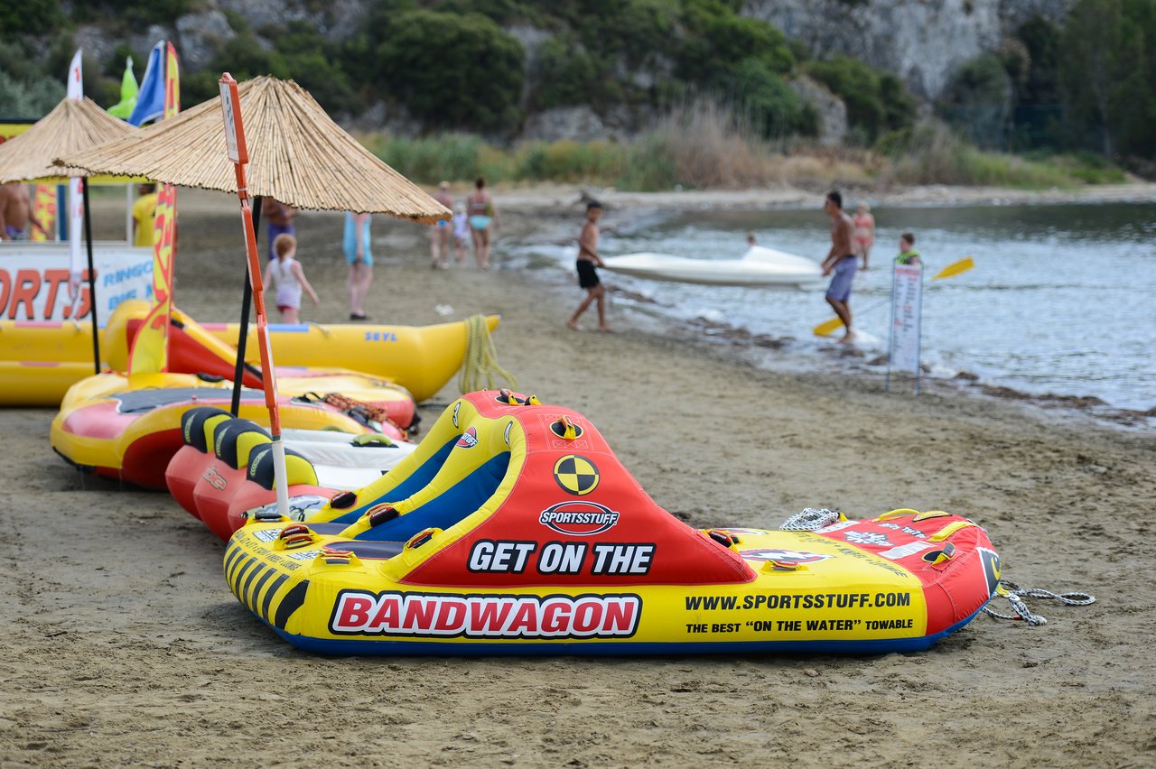 Inflatable water tubes on a sandy beach, with people walking and kayaking near the shoreline.