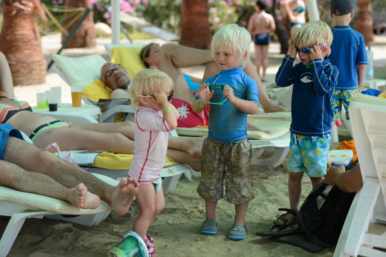 Three young children in swimwear play with sunglasses on a sandy beach while adults relax on lounge chairs nearby.