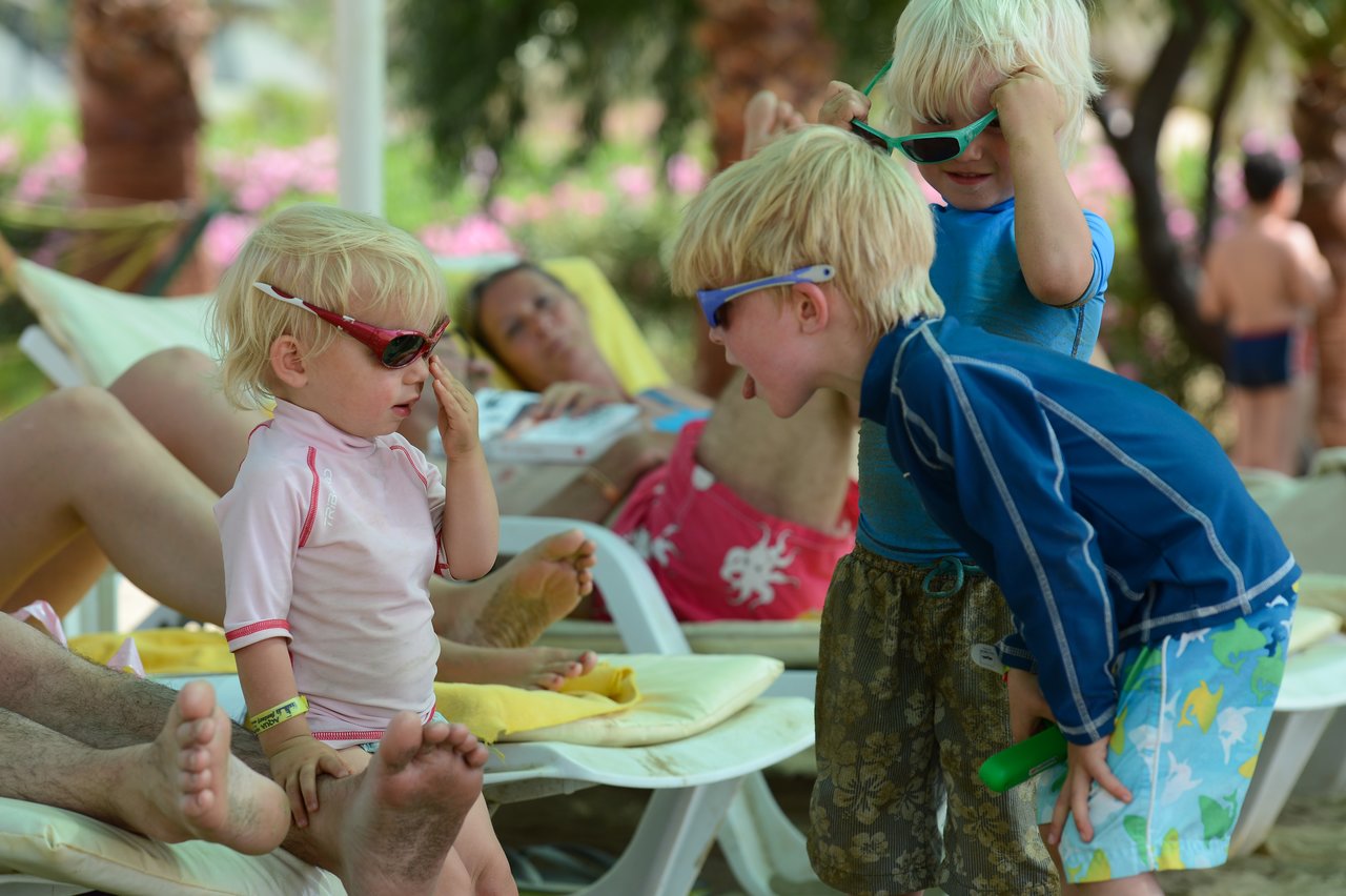 Three children wearing sunglasses interact playfully near lounge chairs, while adults relax in the background.