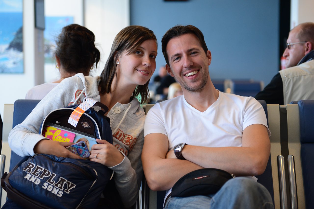 A smiling man and woman sit together in an airport waiting area, with backpacks and travel items beside them.
