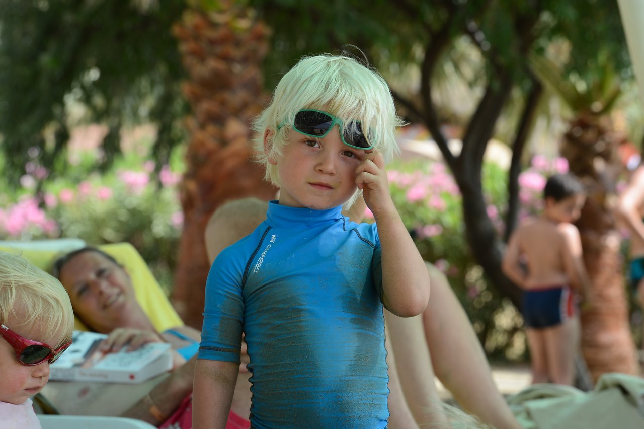 A young child in a blue swimsuit adjusts their sunglasses while standing outdoors near palm trees and other people.