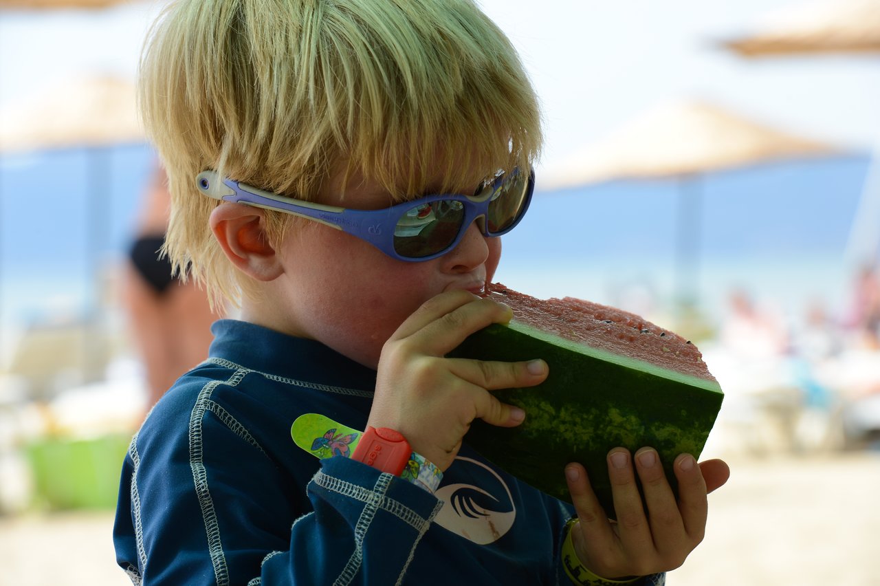 A child wearing sunglasses and a swim shirt eats a large slice of watermelon at the beach.
