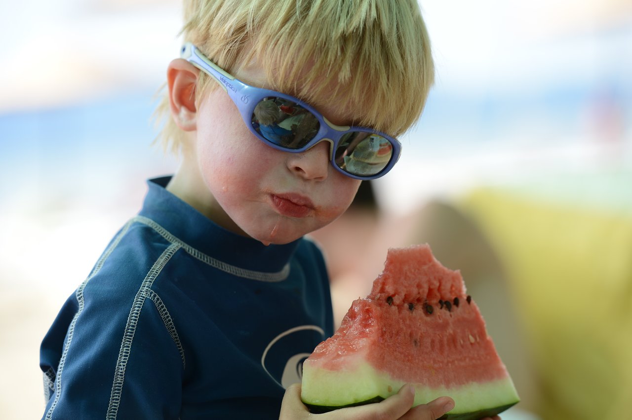 A child wearing sunglasses and a blue shirt eats a large slice of watermelon, with juice dripping down.