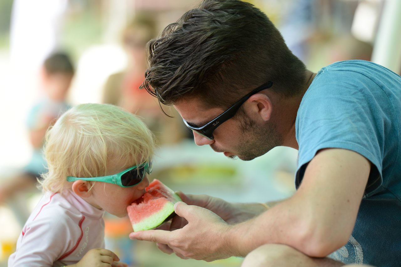 A man holds a slice of watermelon while a young child takes a bite.
