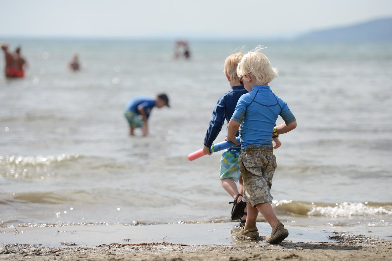 Two children in blue shirts walk along the shoreline, holding toy water guns, while others play in the shallow water.