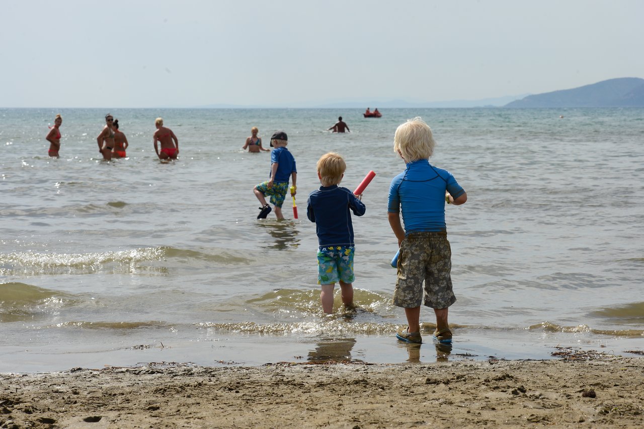 Two young children in swimwear stand at the shore, holding water toys, while others play in the sea.