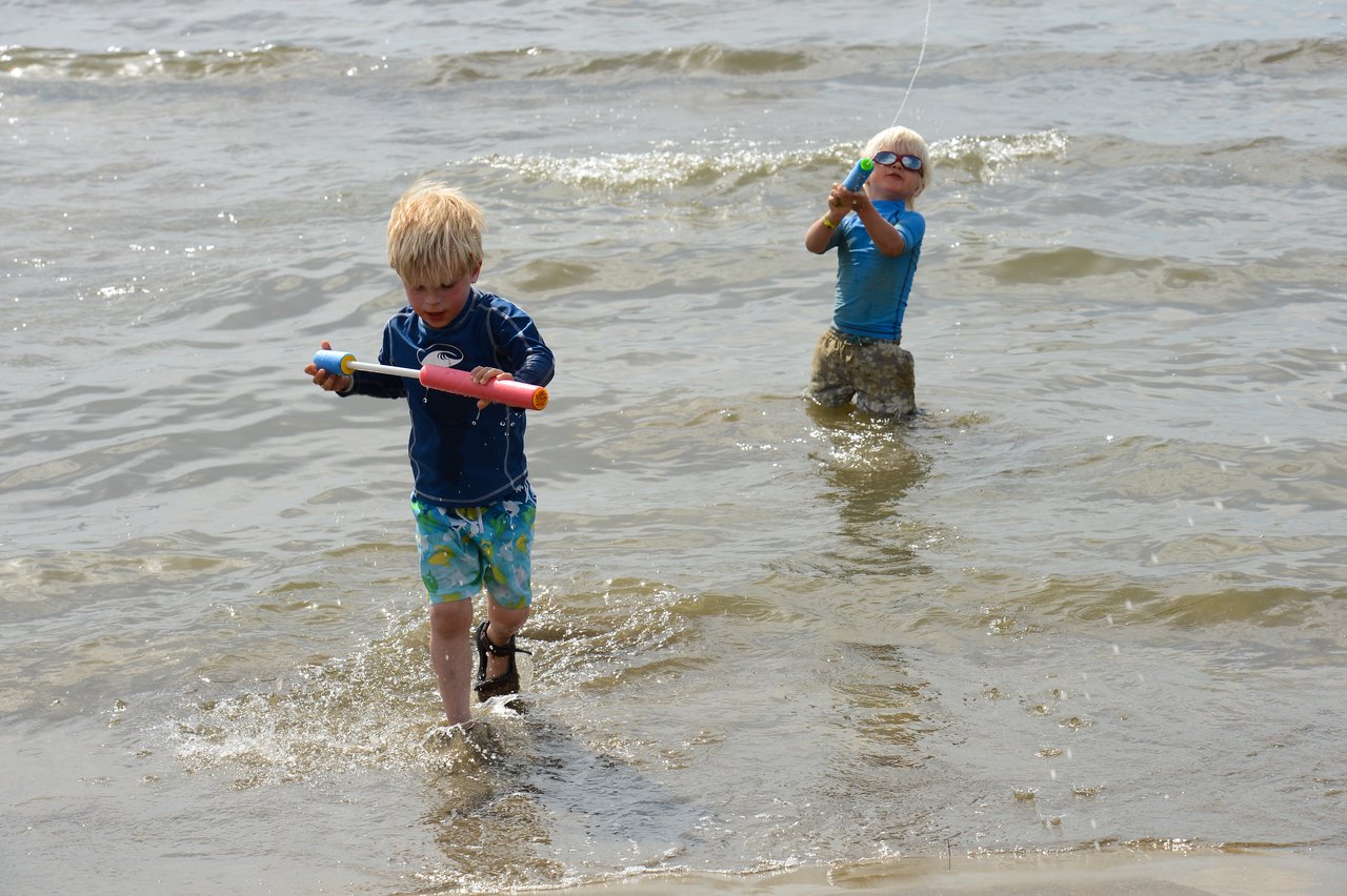 Two children play in shallow water, using toy water guns while splashing around.
