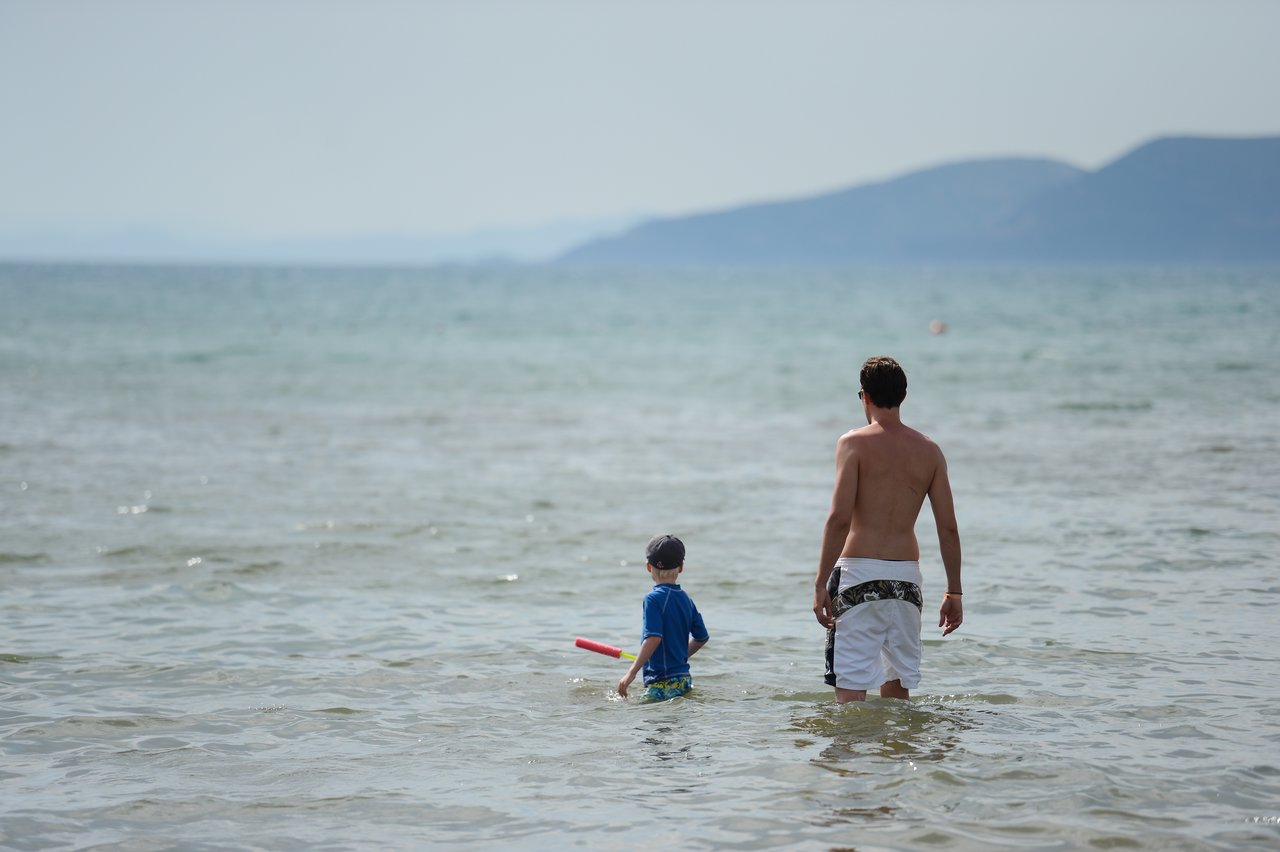A man and a child stand in shallow water at the beach, facing the sea.