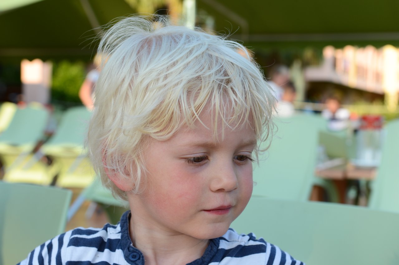 A young child with blonde hair looks down and to the side while sitting outdoors with green chairs around.