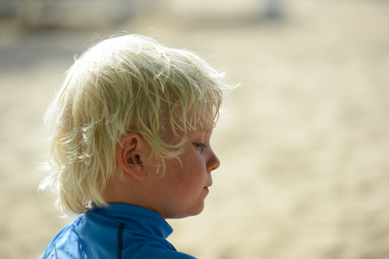 A child with wet blond hair wearing a blue shirt looks down against a blurred sandy background.