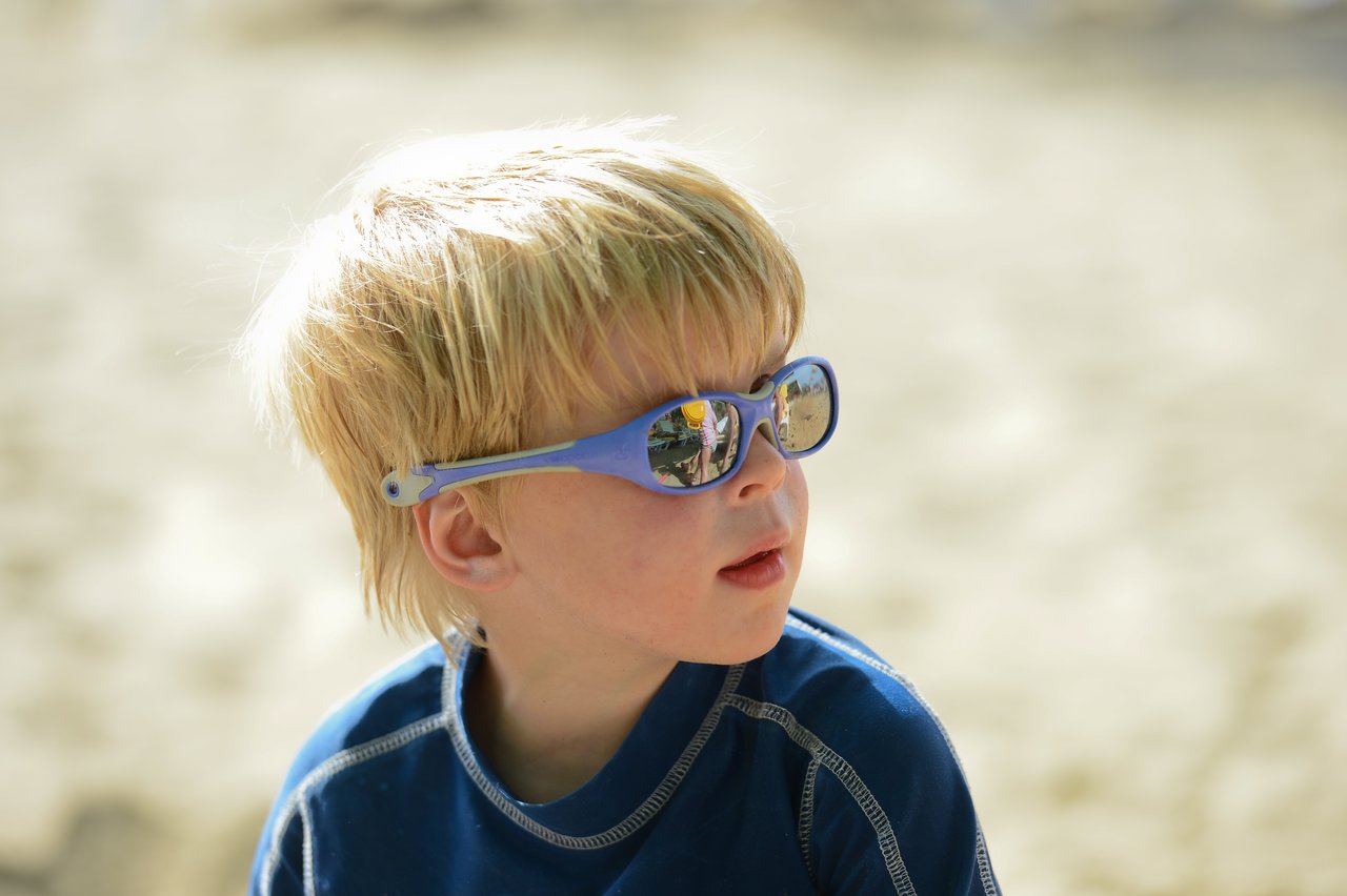 A young child wearing blue sunglasses and a dark shirt looks to the side at the beach.