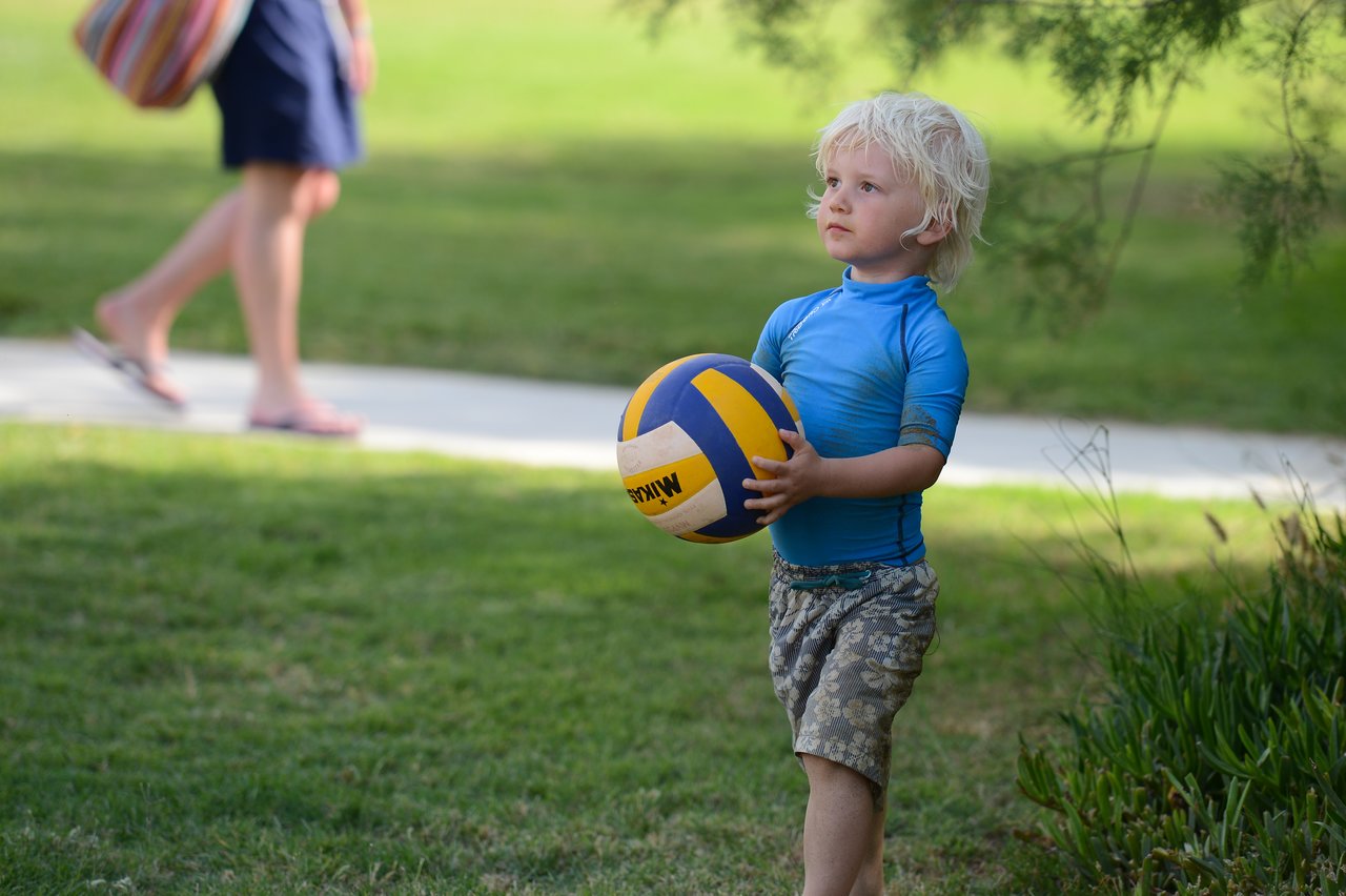 A young child in a blue shirt holds a volleyball while standing on grass, looking to the side.