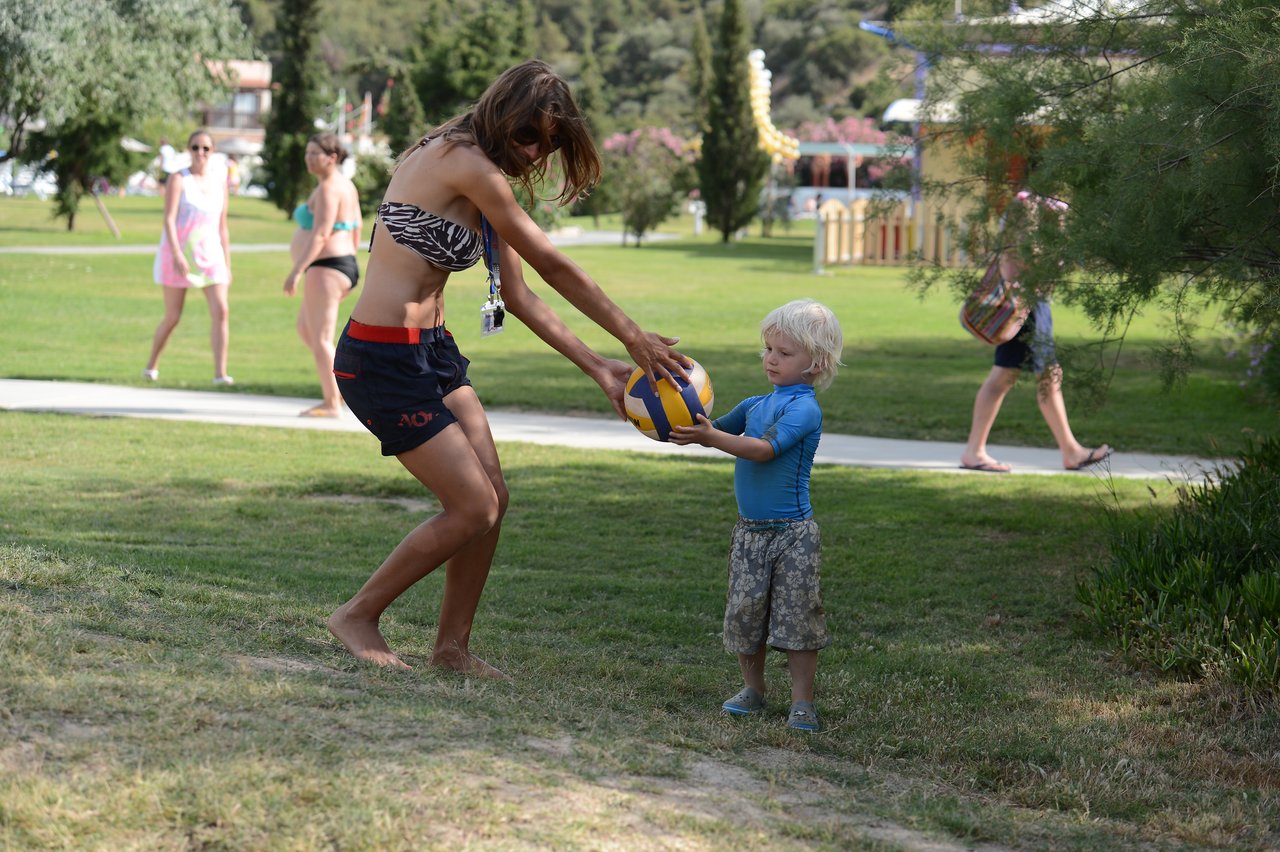 A woman hands a volleyball to a young child in a park with people walking in the background.